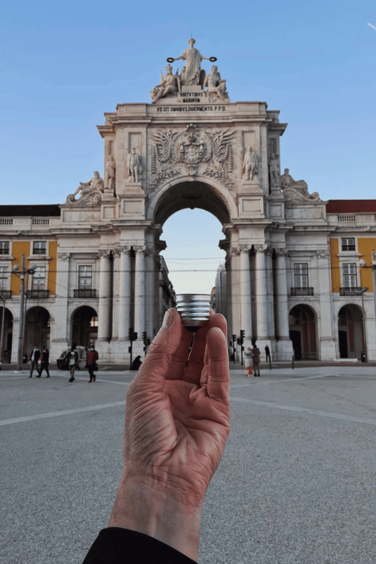 Mano in primo piano che tiene una lampadina davanti a un grande arco monumentale in una piazza, con edifici e passanti sullo sfondo.