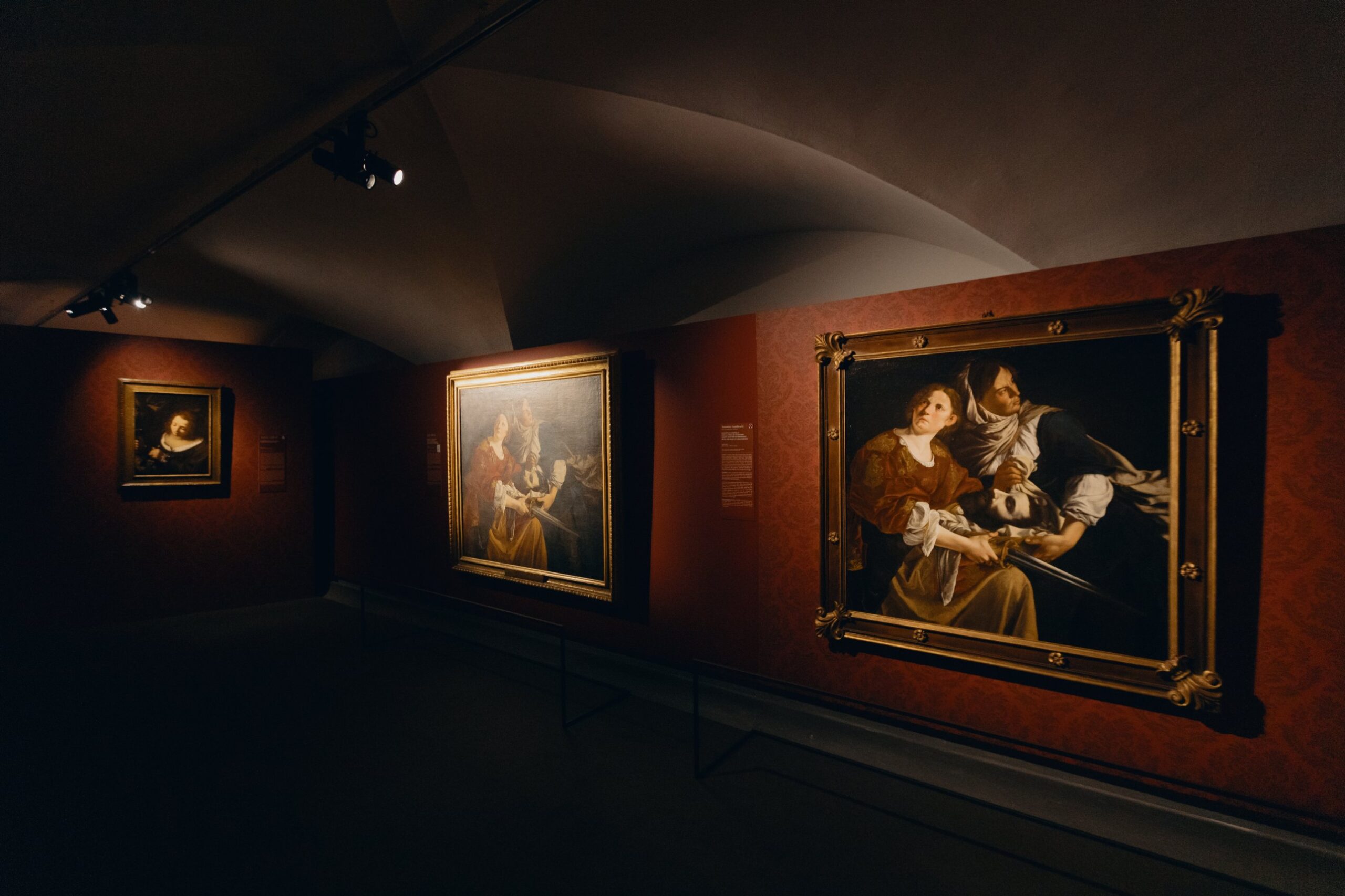 Three paintings by Artemisia Gentileschi on a red damask wall, including *Judith and the Maidservant with the Head of Holofernes*.