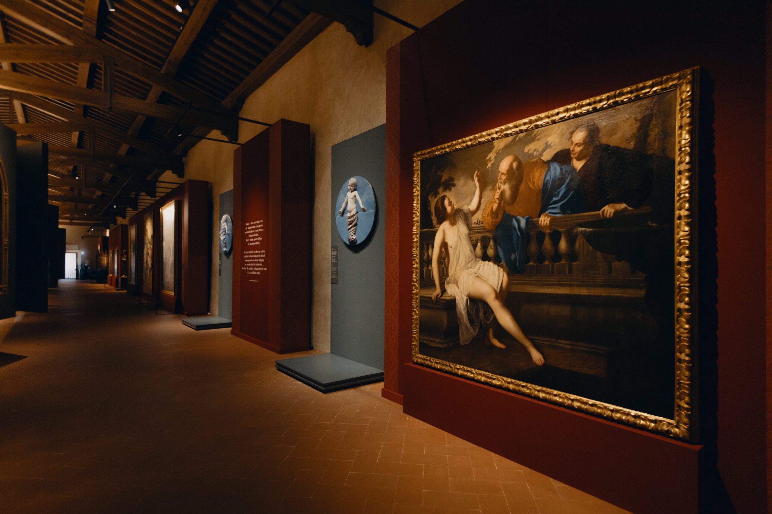 A gallery with a wooden-beamed ceiling, the large painting *Susanna and the Elders*, and a Robbiano tondo depicting a baby in swaddling clothes.
